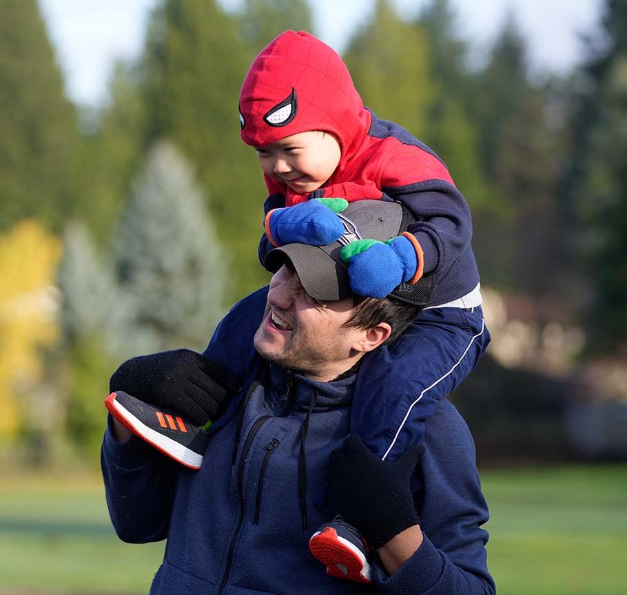 father in baseball cap carries boy in Spiderman costume on his shoulders