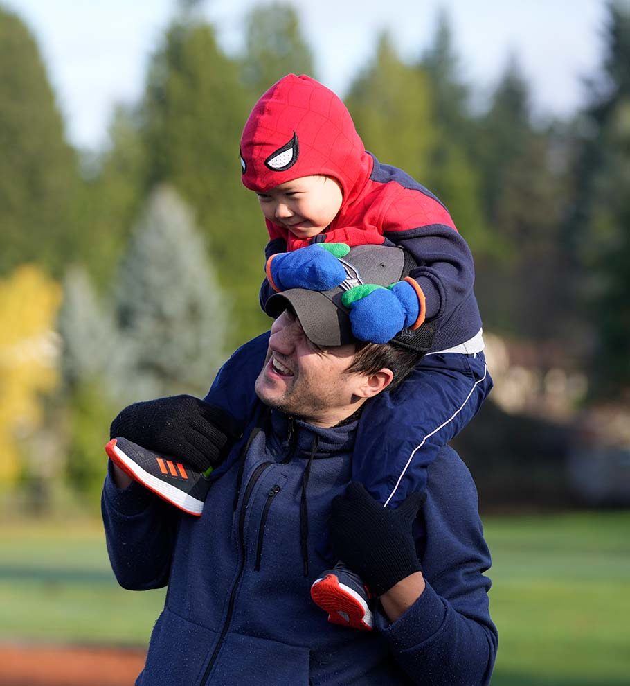 father in baseball cap carries boy in Spiderman costume on his shoulders