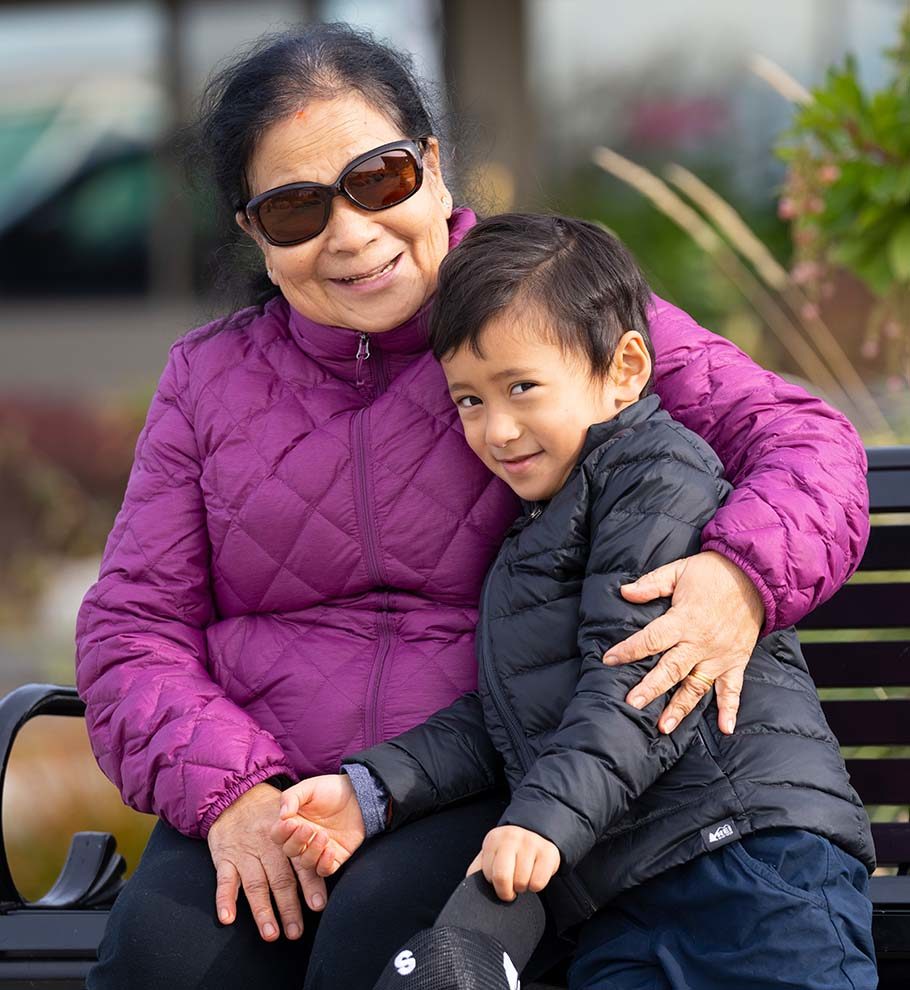 grandmother in sunglasses and magenta down coat sitting on park bench hugs smiling grandson