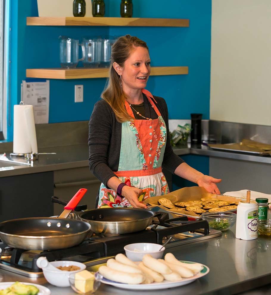 woman in bright orange and blue apron leads cooking class in test kitchen