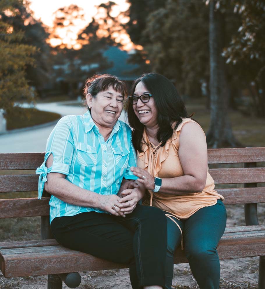 two hispanic women sitting on a bench leaning in towards each other and laughing